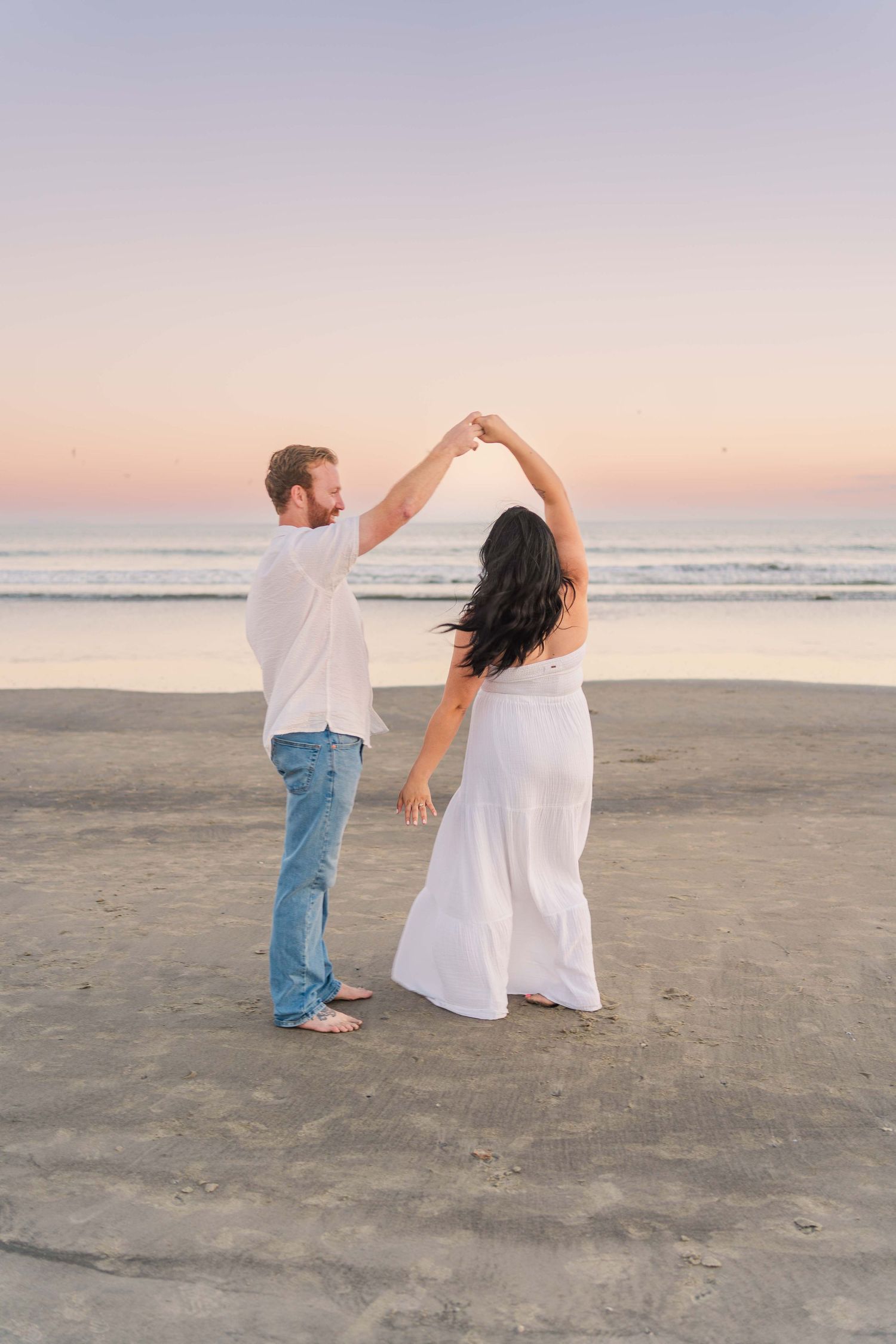 A couple in white clothing dances together on a beach at sunset with ocean waves in the background.