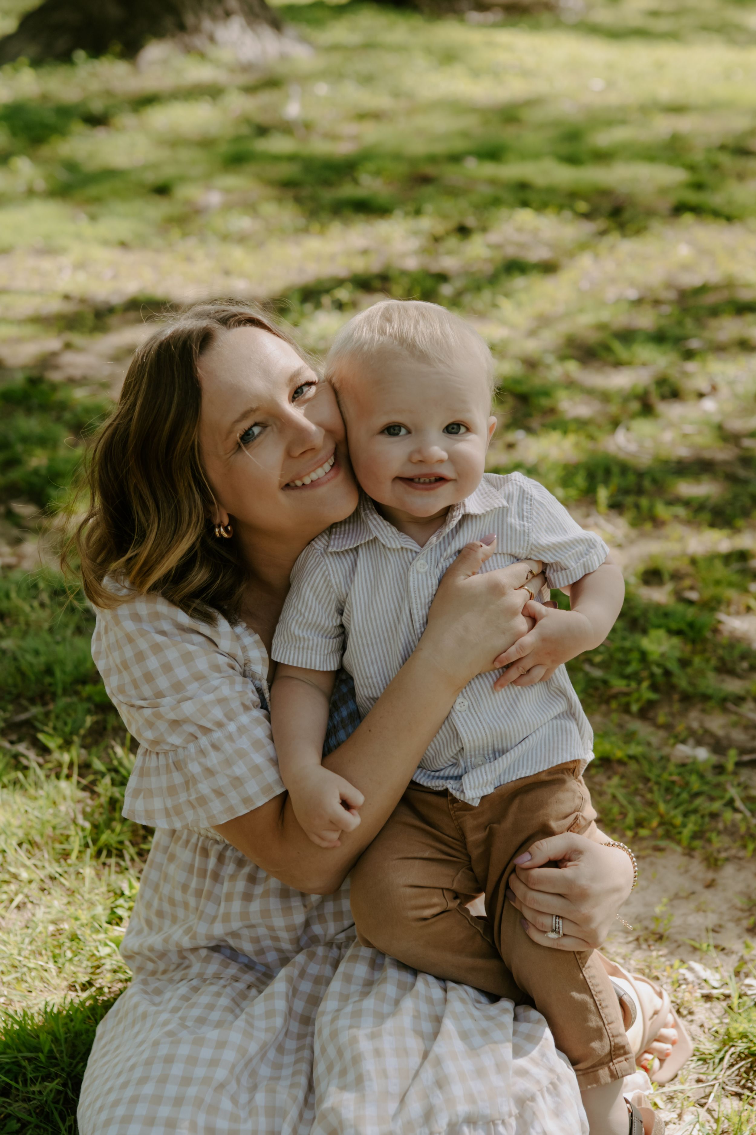 Tender family moment shared on grass in sunlit park setting.