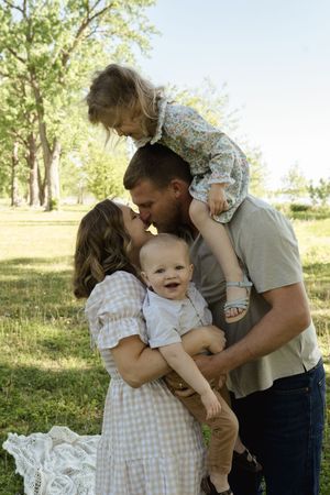 A tender family moment captured in a sun-dappled outdoor setting with natural lighting and greenery.