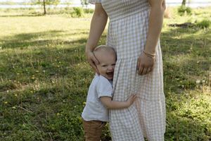 A small child in light clothing holds onto a gingham dress in dappled sunlight.