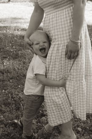 A black and white capture of a laughing toddler hugging a gingham dress outdoors.