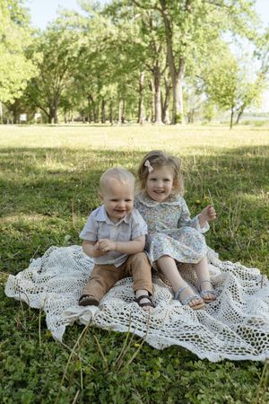 Children sitting on lace blanket in sunny park with trees in background.