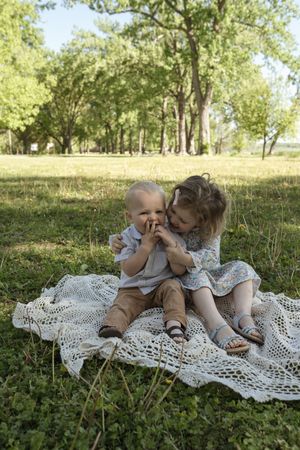 Two children sitting together on a white lace blanket in a sunny park with trees in the background.