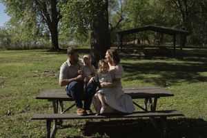 A family sits together on a wooden picnic bench in a park setting.