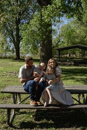 A family cuddles close together on a park picnic bench surrounded by green trees.
