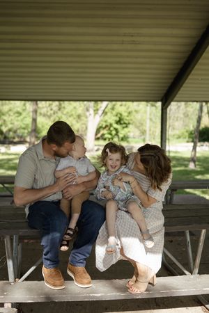 A family shares a playful moment together on a wooden bench in a shaded outdoor setting.