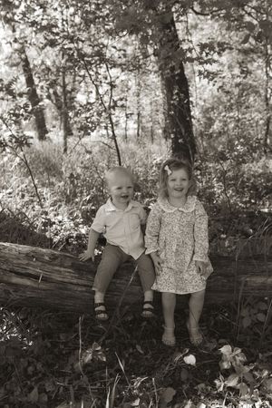 Black and white portraits of children sitting together on rustic log in natural woodland setting.