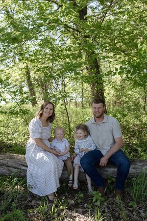 A casual family portrait session on a log in a wooded area with dappled sunlight.