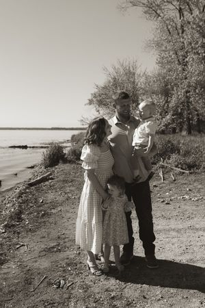 A black and white portrait of a family group standing together near bushes by the water.