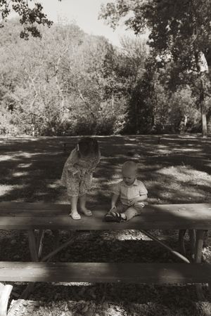 Black and white photo of children sitting on a wooden picnic table in a shaded park.