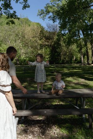 Family gathered around a wooden picnic table in a sunny park setting with lush trees.
