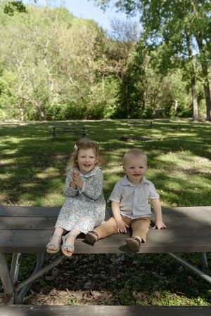 Two young children sitting together on a wooden bench in a sunlit park.