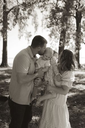 A playful family moment captured in black and white under trees in a park setting.
