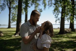A romantic couple embraces under tall trees in a sunlit outdoor setting.