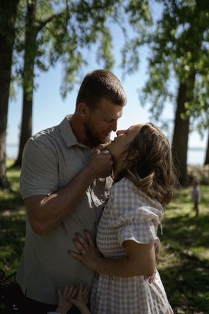 A couple shares a romantic embrace in a serene outdoor setting with sunlit trees.