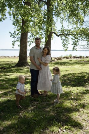 A family group interacts playfully in a sunny park setting with trees in the background.