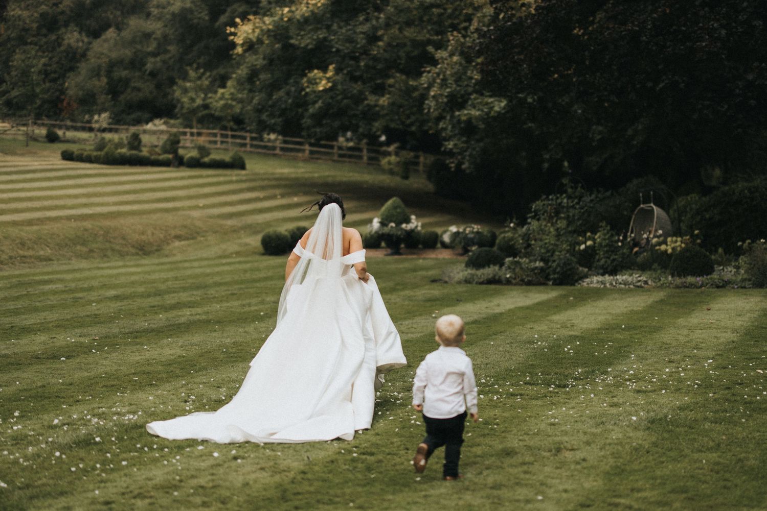 A bride in a flowing white gown walks across a manicured lawn with a small child in formal attire.