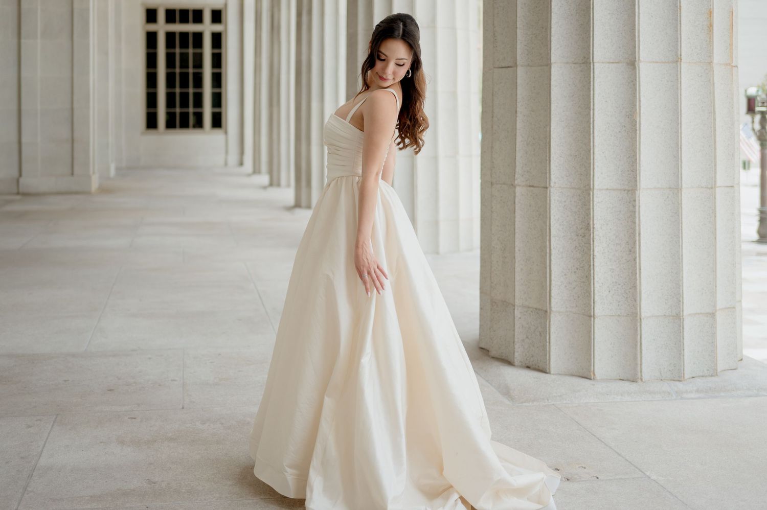 Elegant bride in flowing ivory gown poses gracefully between white marble columns in a classic architectural setting.