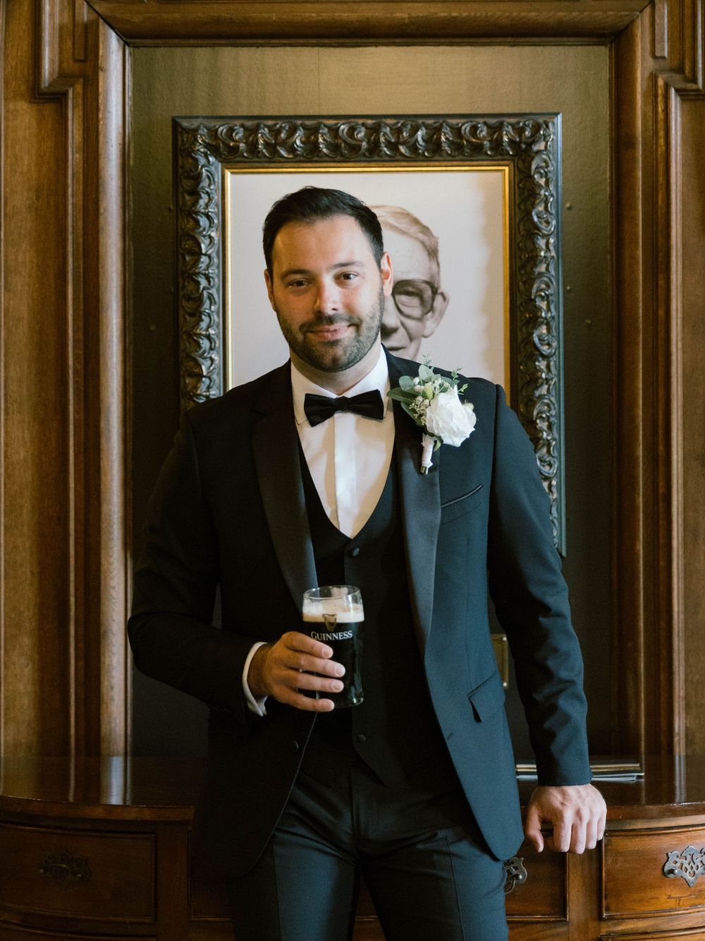 A formal wedding party member in a black tuxedo holds a dark beer while posing in front of an ornate framed mirror.