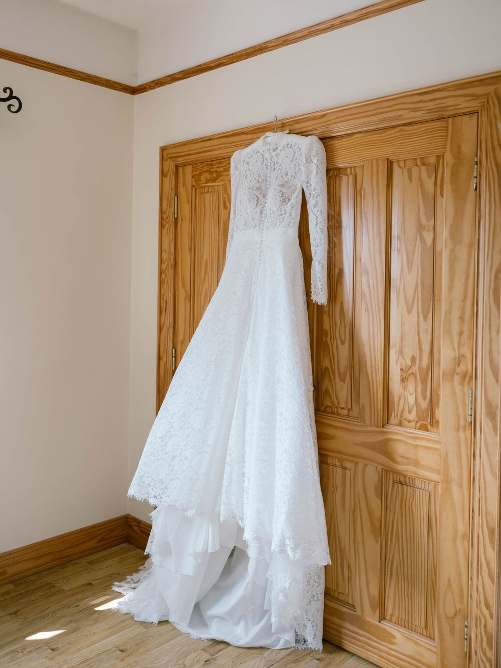 A white lace wedding dress with long sleeves hanging on a wooden door in natural light.