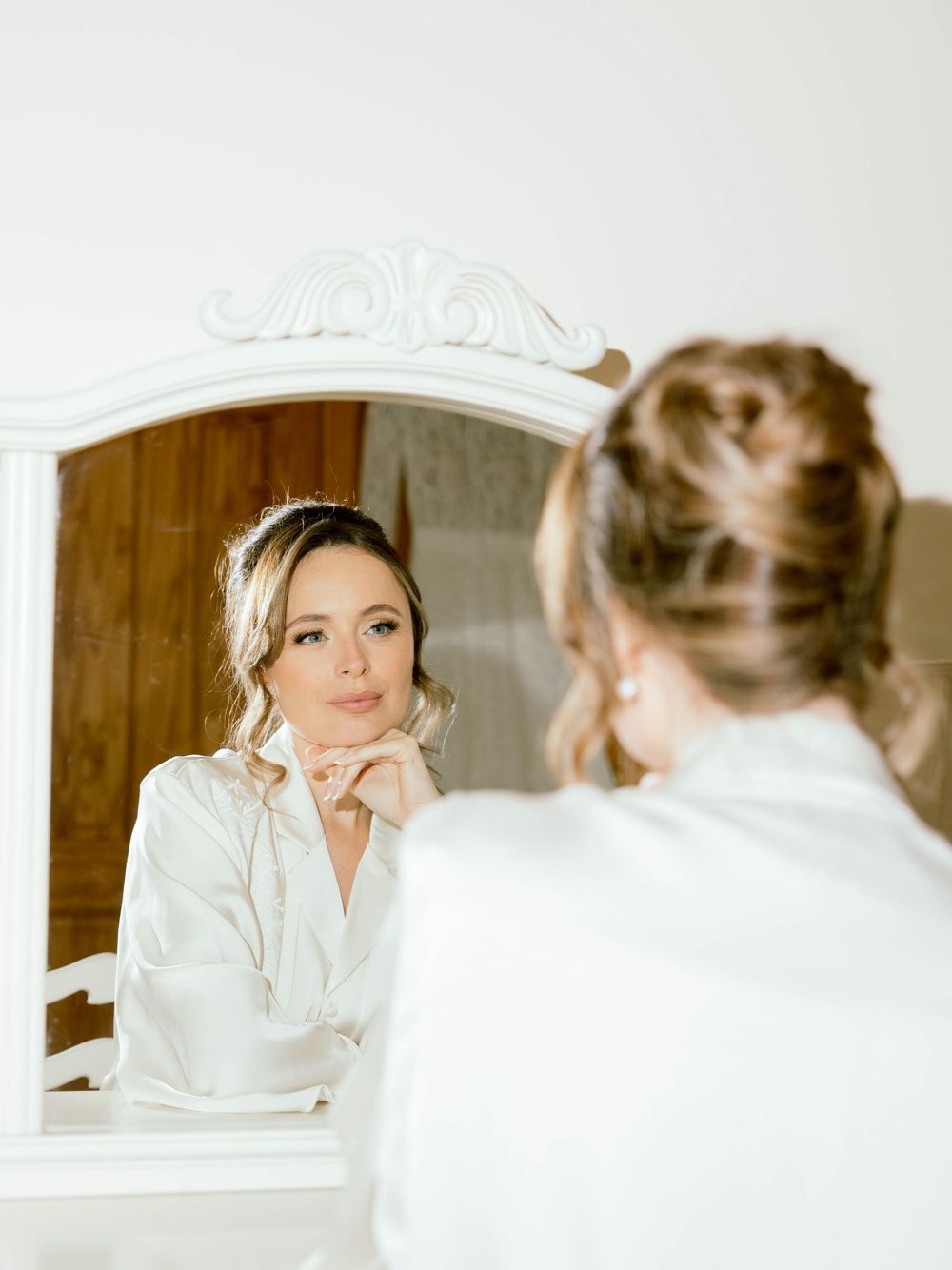 A bride in a white robe looks at her reflection in an elegant vanity mirror during wedding preparations.