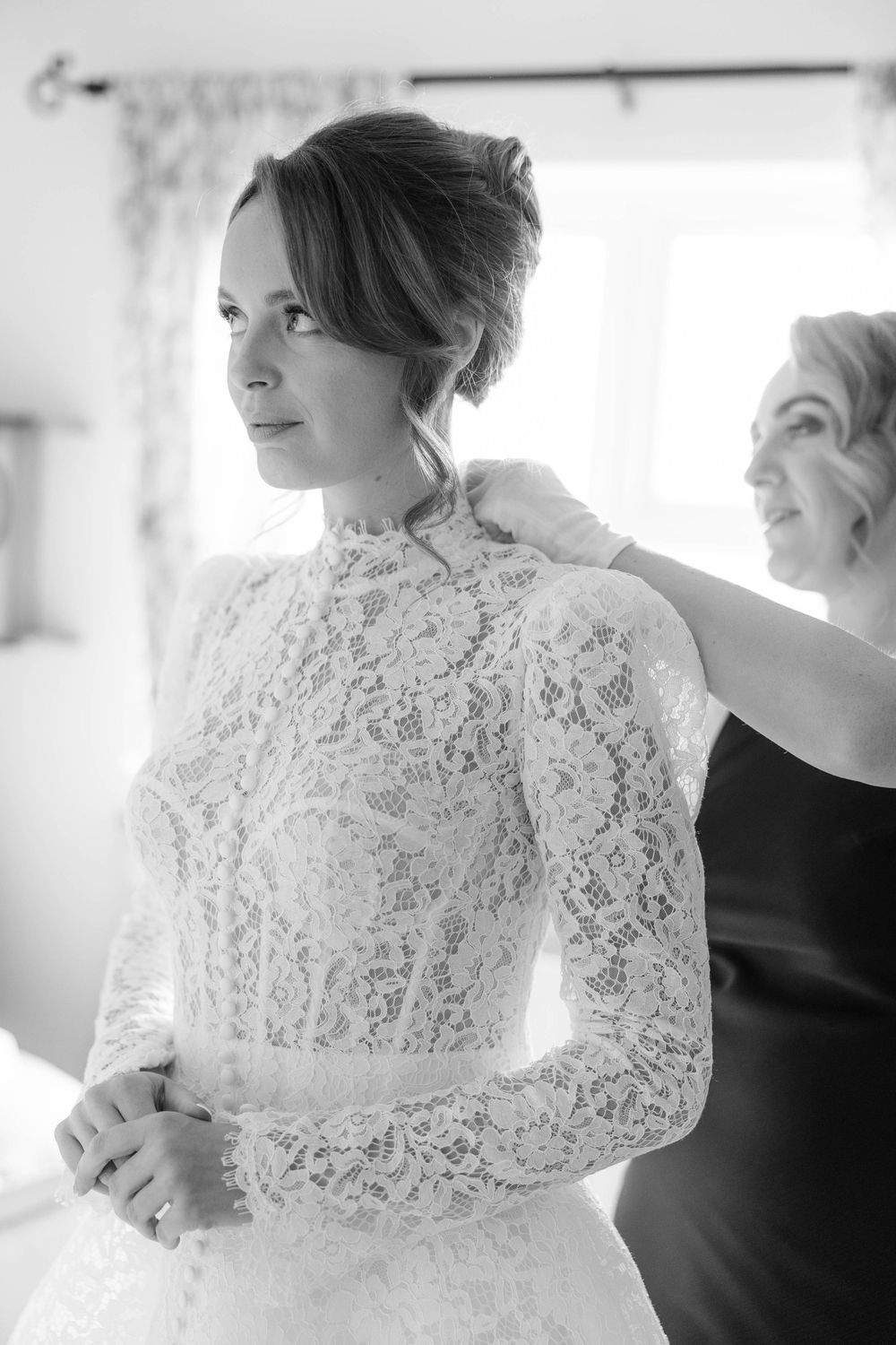 A delicate lace wedding dress with long sleeves being fitted in a bright room.