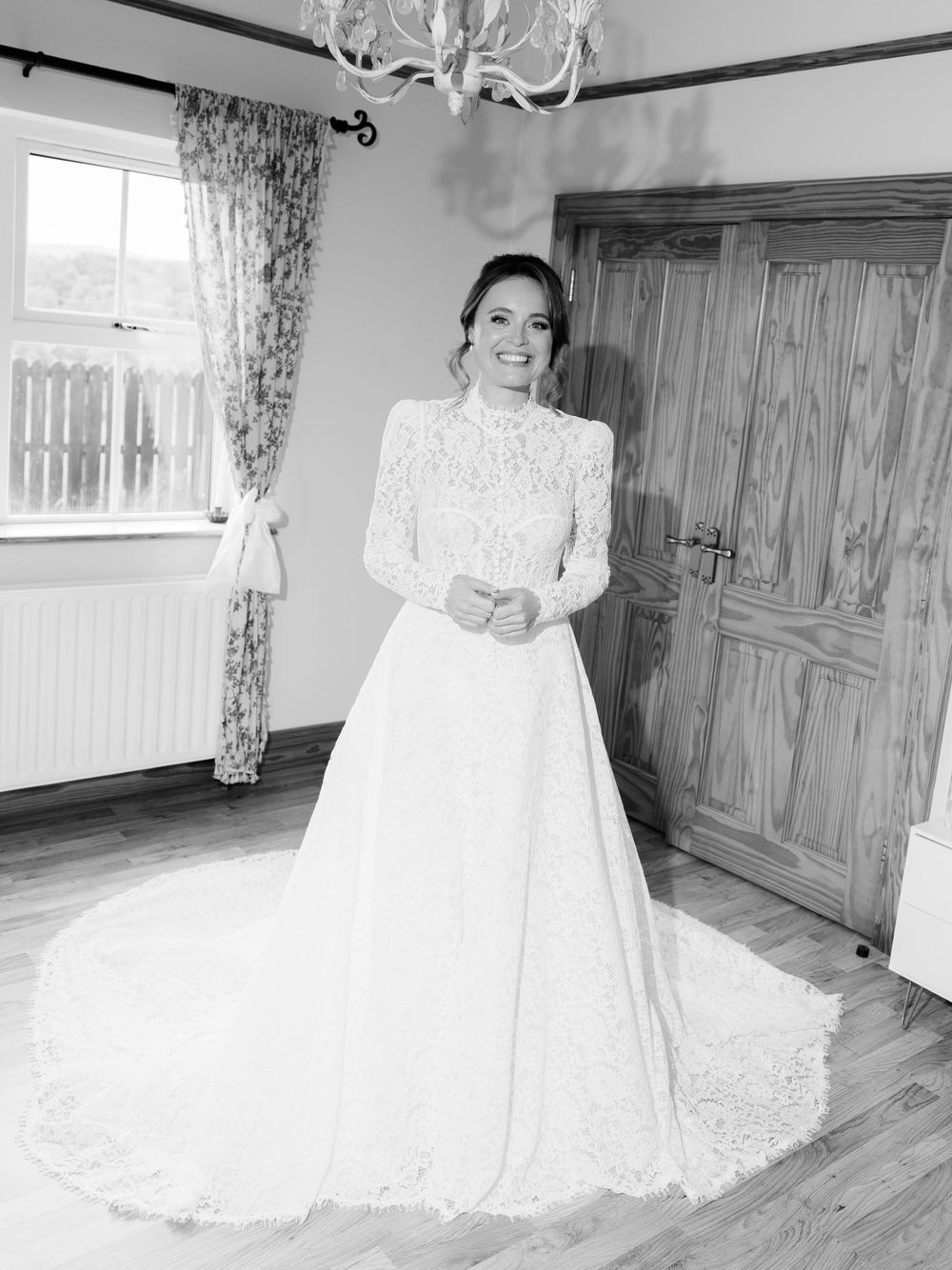 A bride in a long-sleeved lace wedding gown stands in a rustic room with wooden floors and a chandelier.