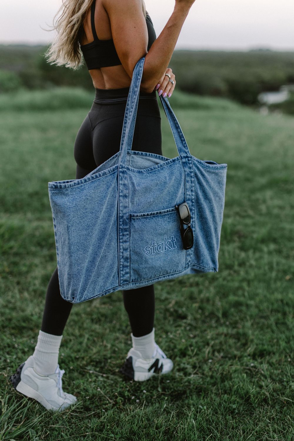 Woman in black athleisure holding a large light wash denim tote bag outdoors on green grass.