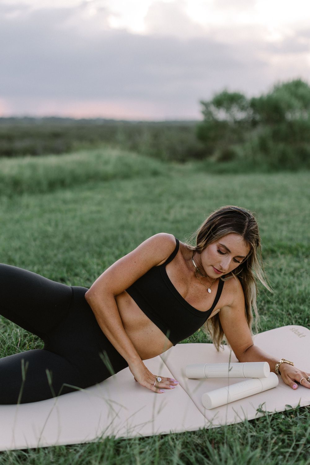 Woman in black activewear practicing yoga on a mat outdoors at sunset in a lush green field.