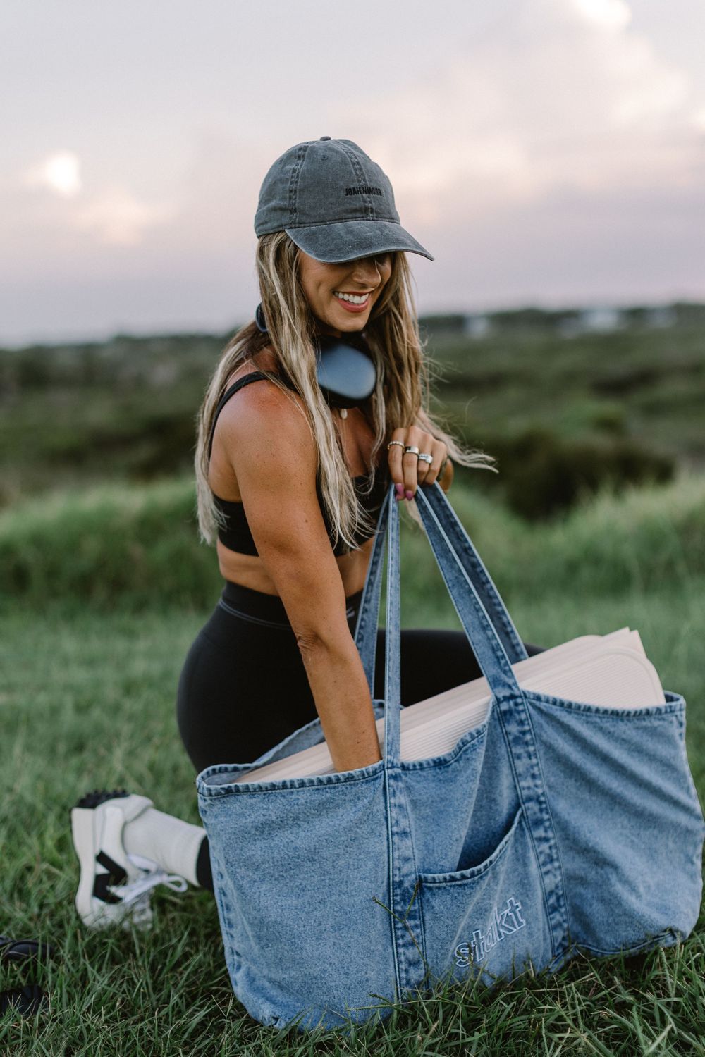 Woman in black sports bra and leggings smiling while holding a large denim tote bag outdoors at sunset.
