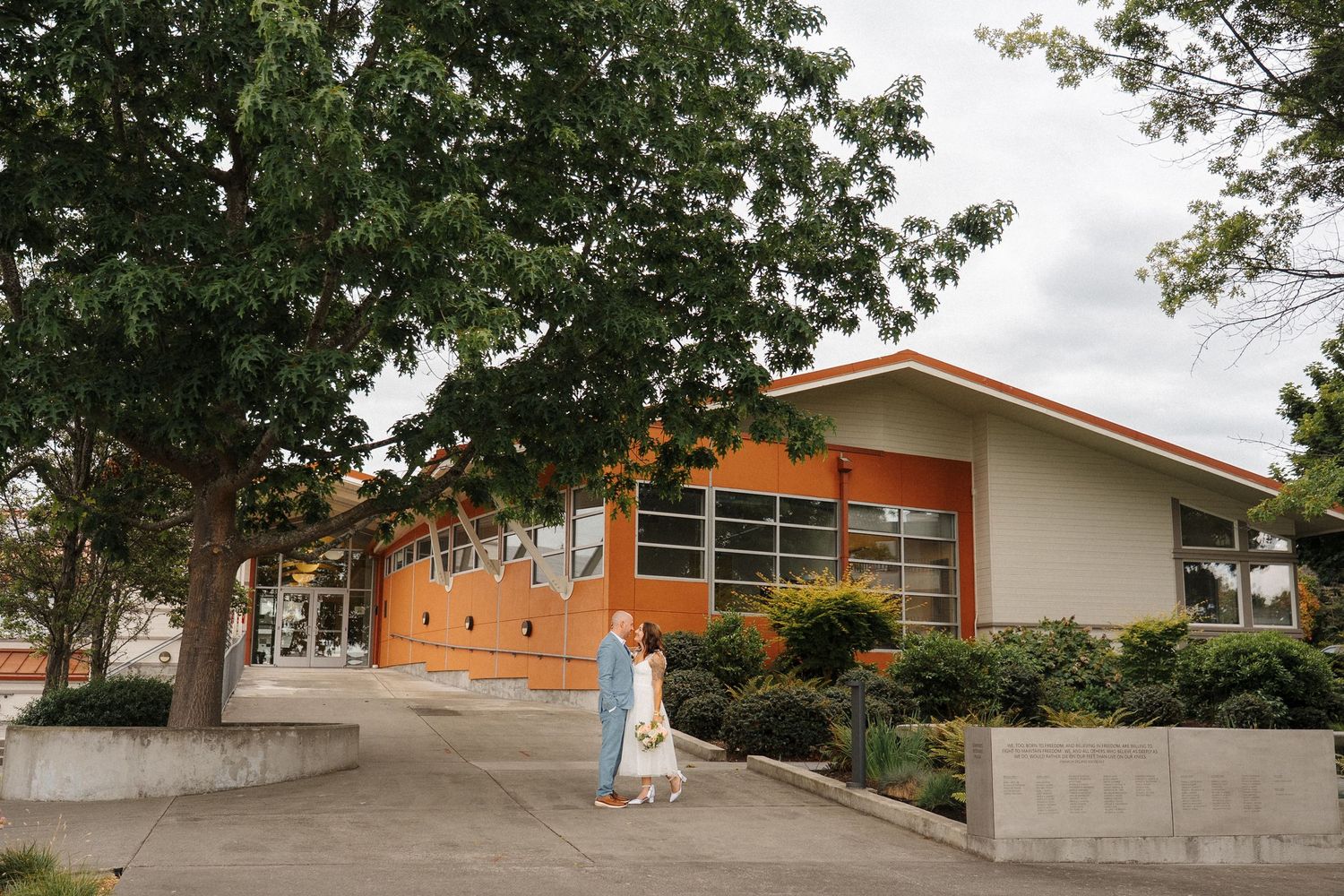 A couple stands outside a modern orange building with slanted roof and concrete retaining walls surrounded by trees.