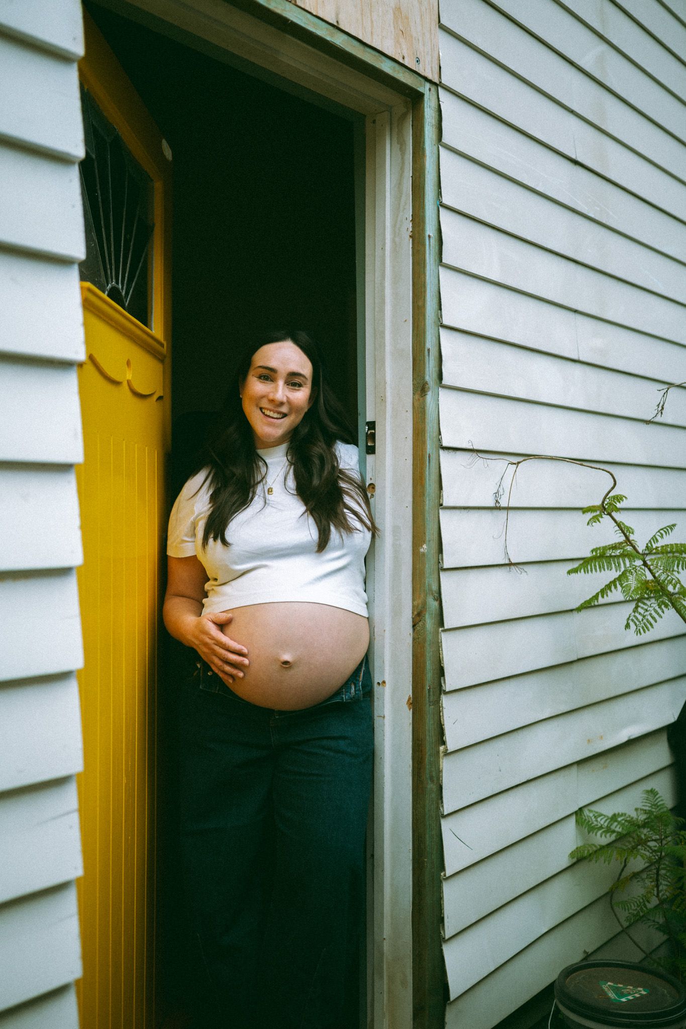 A parent in an off-shoulder brown dress holds a sleeping baby wrapped in a white blanket near a staircase.