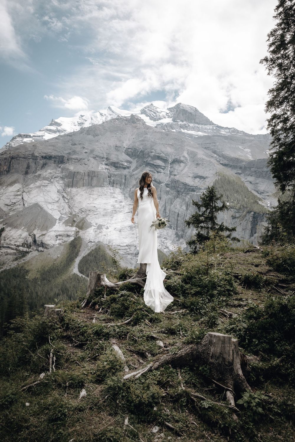 A white-dressed figure stands on a mountain slope against snow-capped peaks in a dramatic alpine landscape.