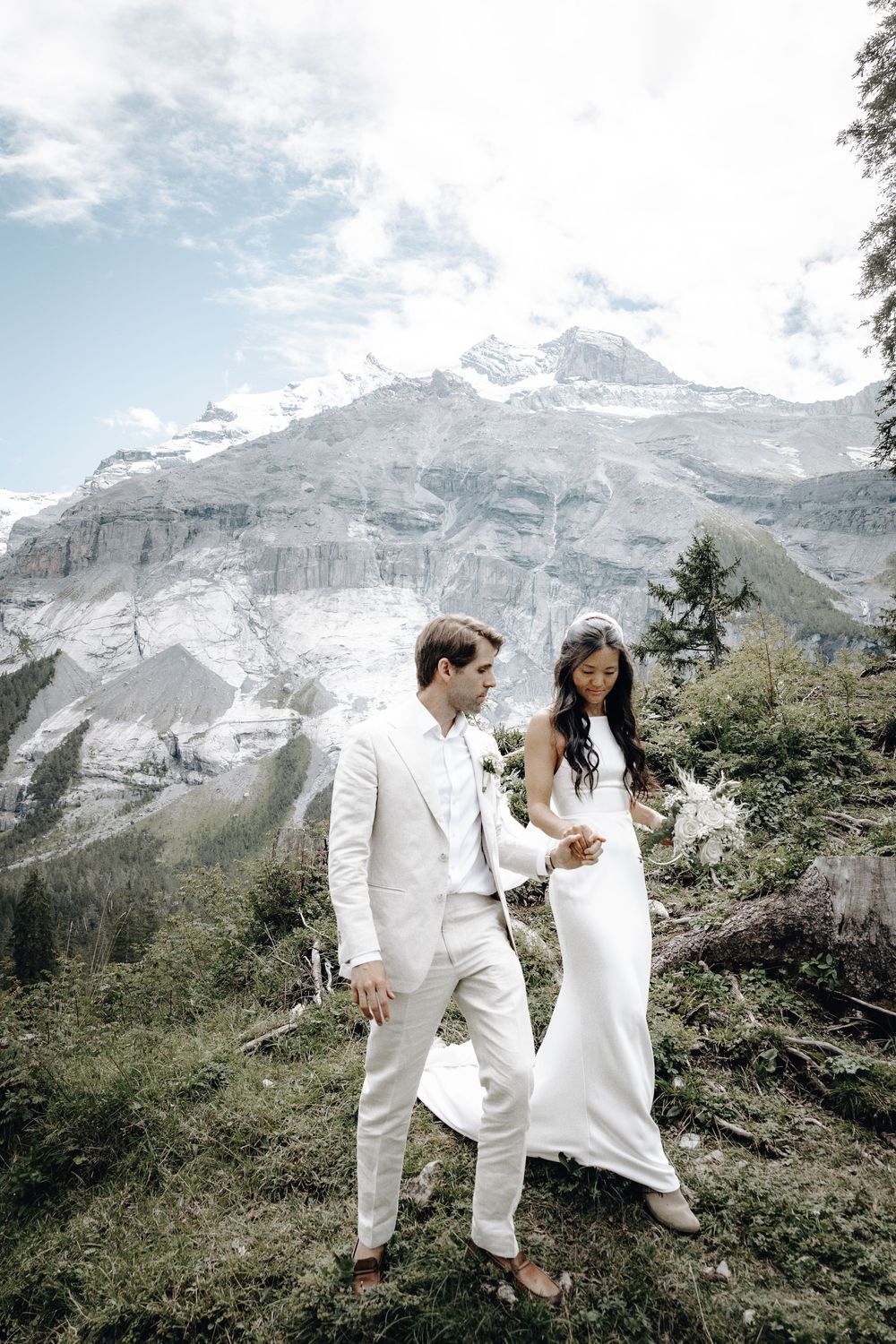 A couple in white attire shares a romantic moment against a stunning mountain backdrop with snow-capped peaks.