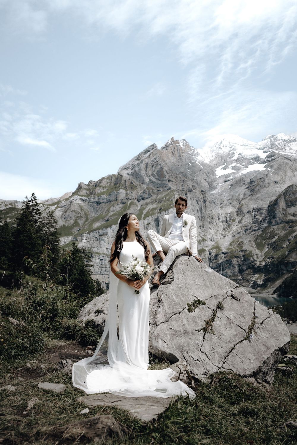Couple in white stands on rocky outcrop with dramatic snow-covered mountain peaks in background.
