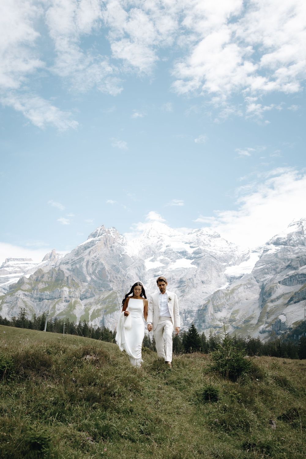 A couple in all-white outfits walk along a mountain ridge with snow-capped peaks in the background.