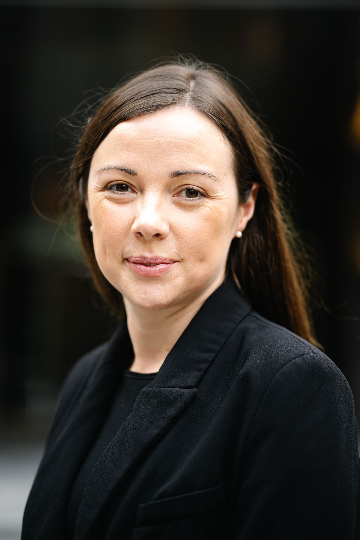 Professional headshot of a woman with dark hair wearing a black blazer outdoors.