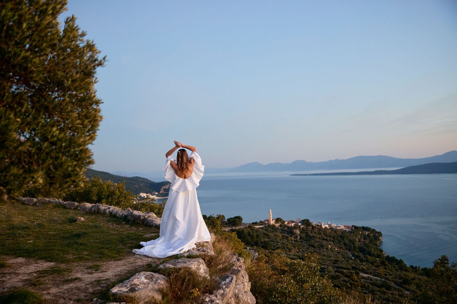 A person in white stands on coastal rocks overlooking a calm sea and distant mountains.