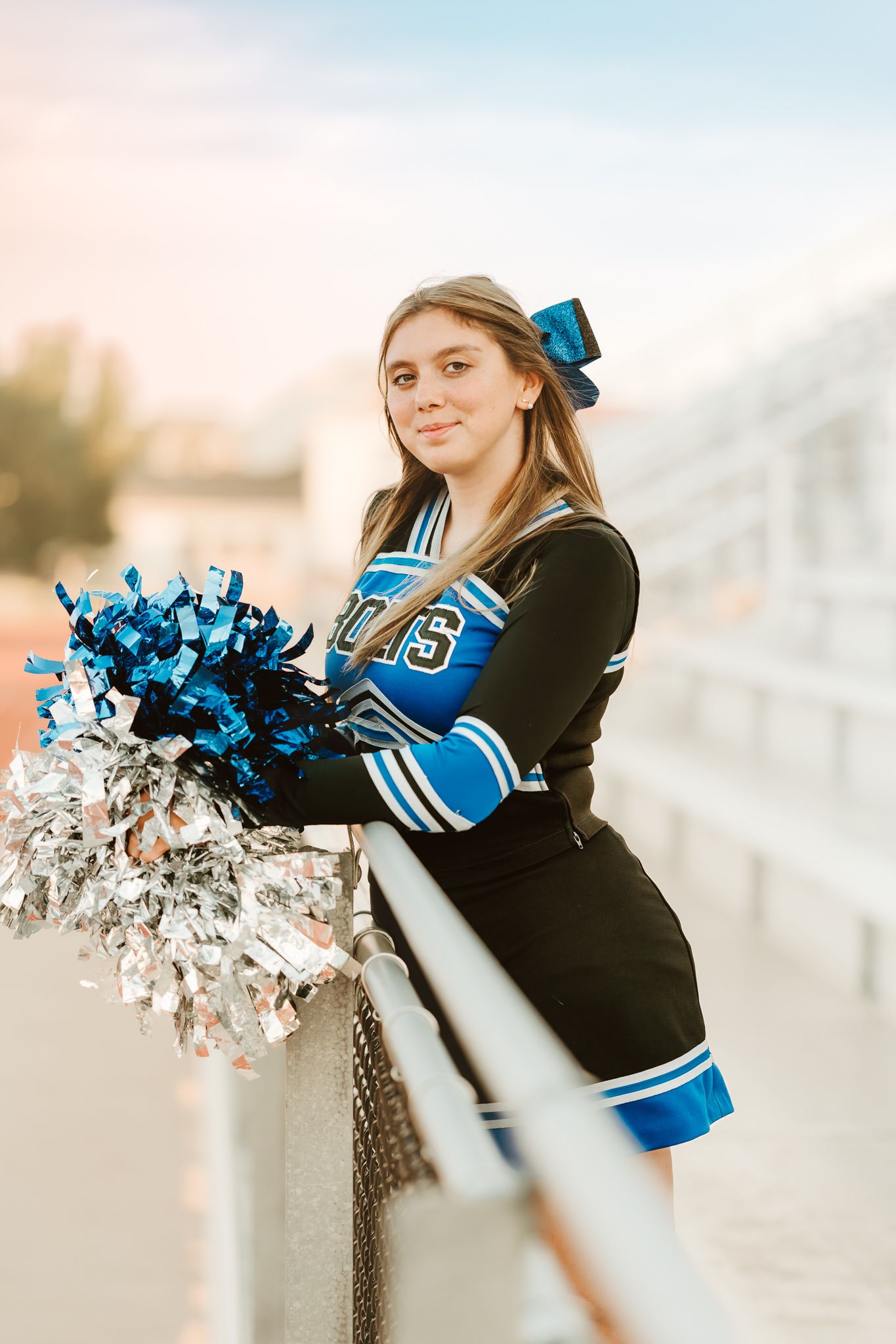 Cheerleader in blue and black uniform holds pom poms during sunset performance.