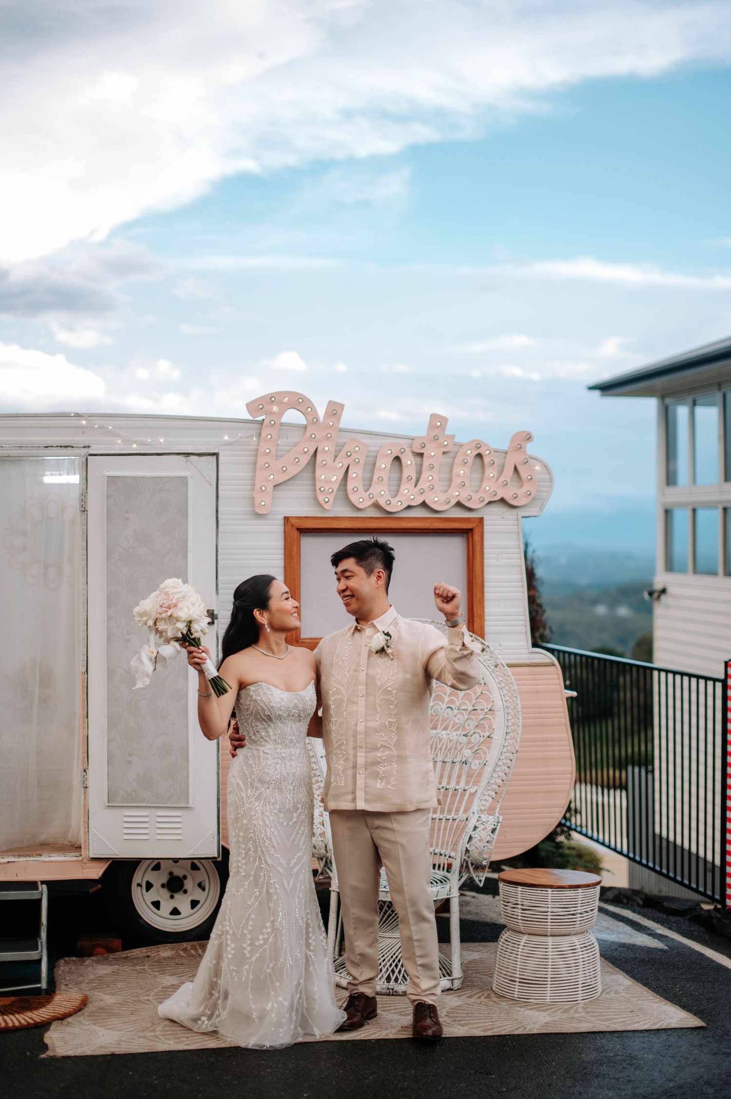 Wedding couple standing by a retro photo booth trailer with decorative wings at an outdoor venue.