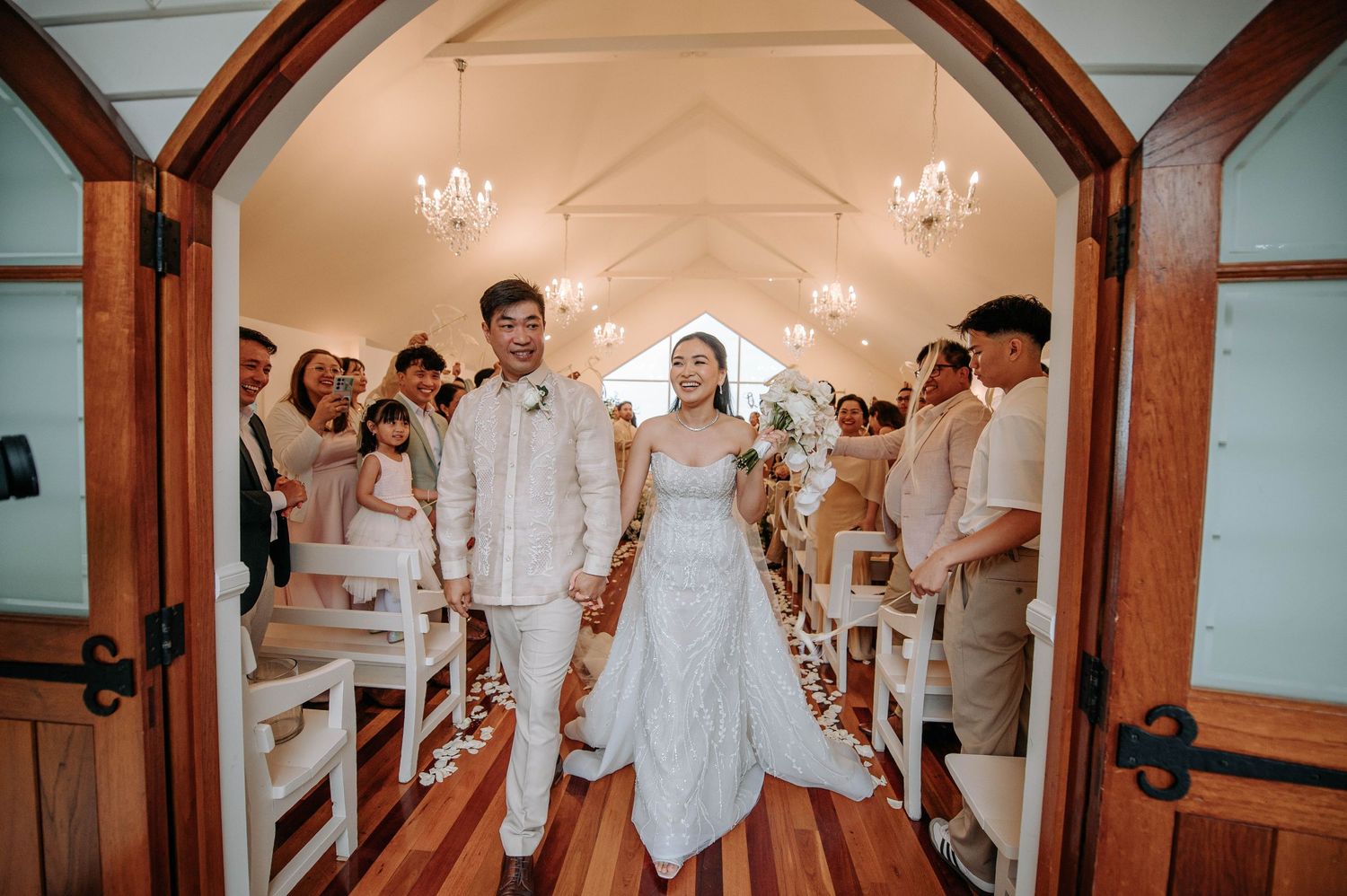 A newlywed couple exits through rustic wooden church doors while guests look on.