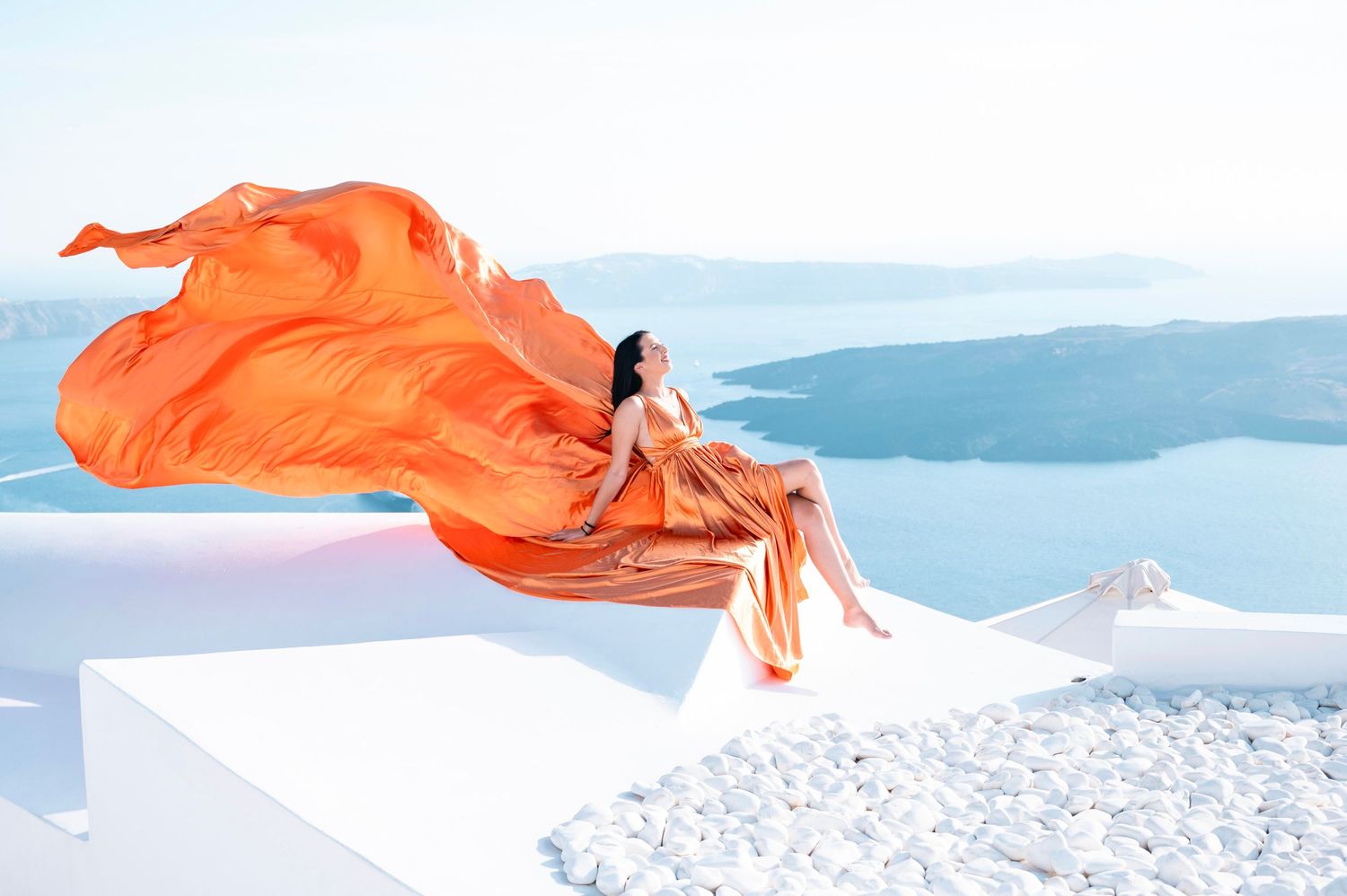 Woman in flowing orange dress sitting on white wall overlooking Mediterranean Sea in Santorini.