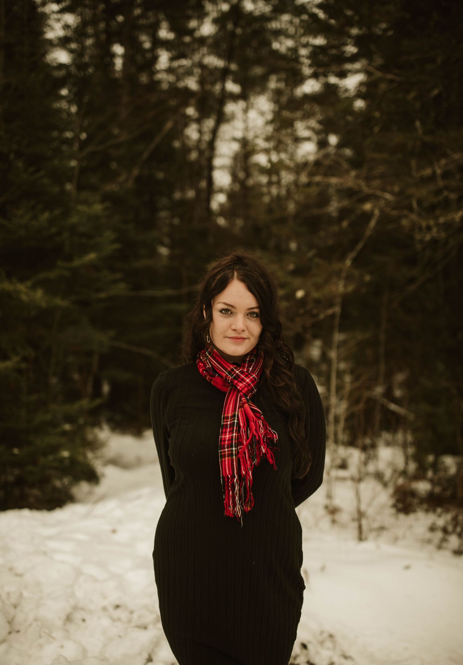 A winter portrait of a woman in black wearing a red plaid scarf in a snowy forest.