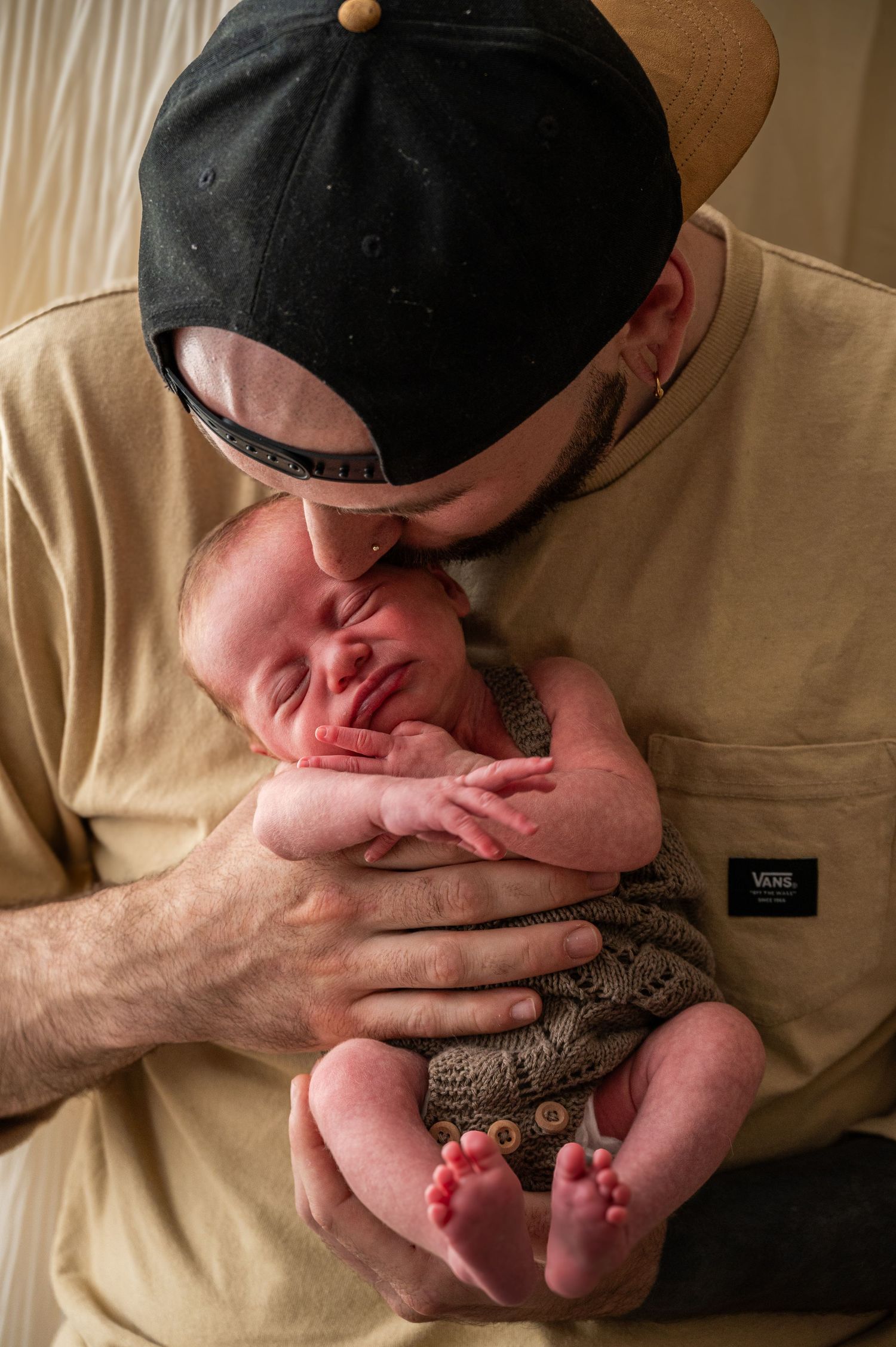 Father in baseball cap tenderly holding newborn baby wearing knit clothing.