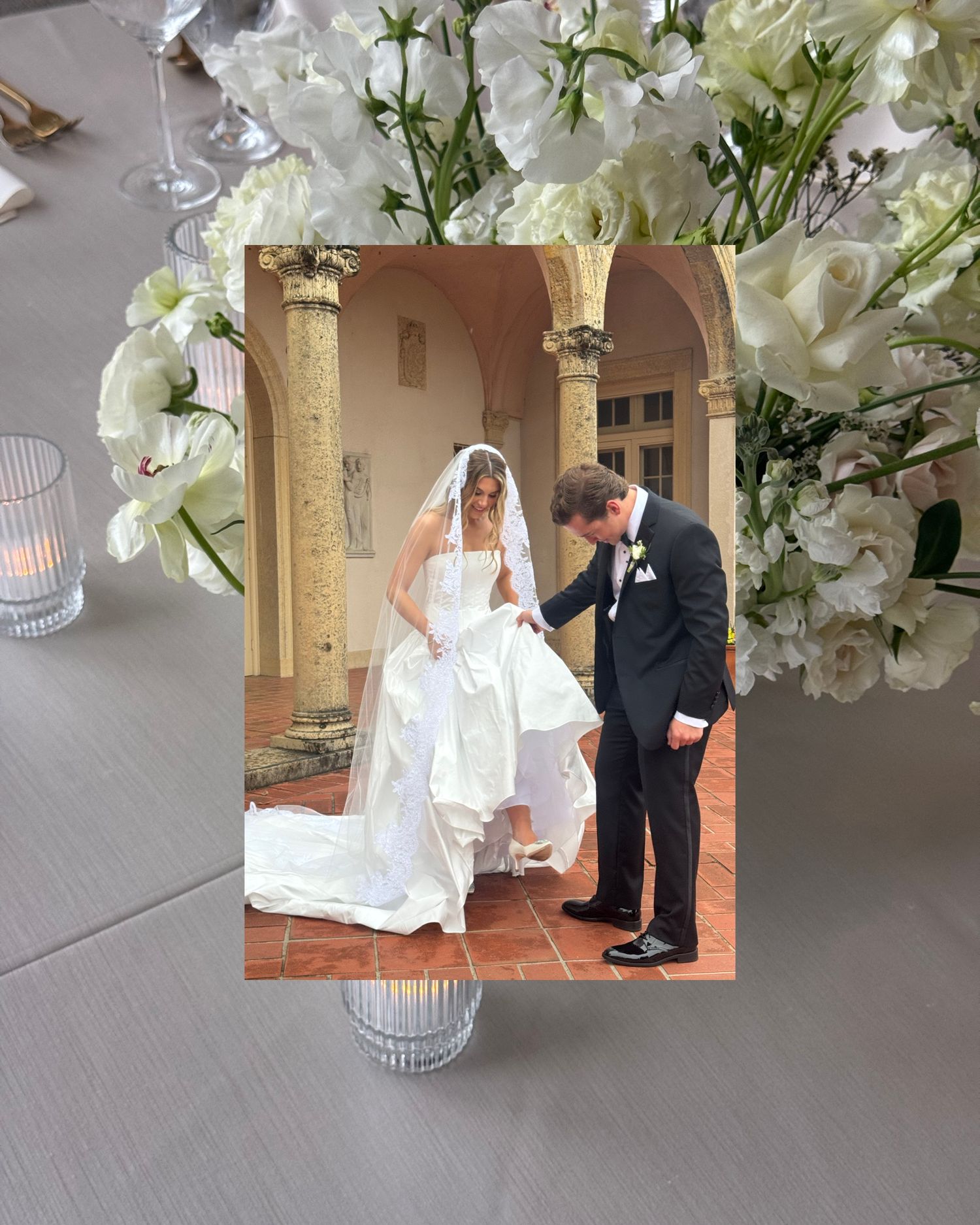A wedding photo displayed in a white floral centerpiece with mercury glass votive candle holders on a gray linen tablecloth.