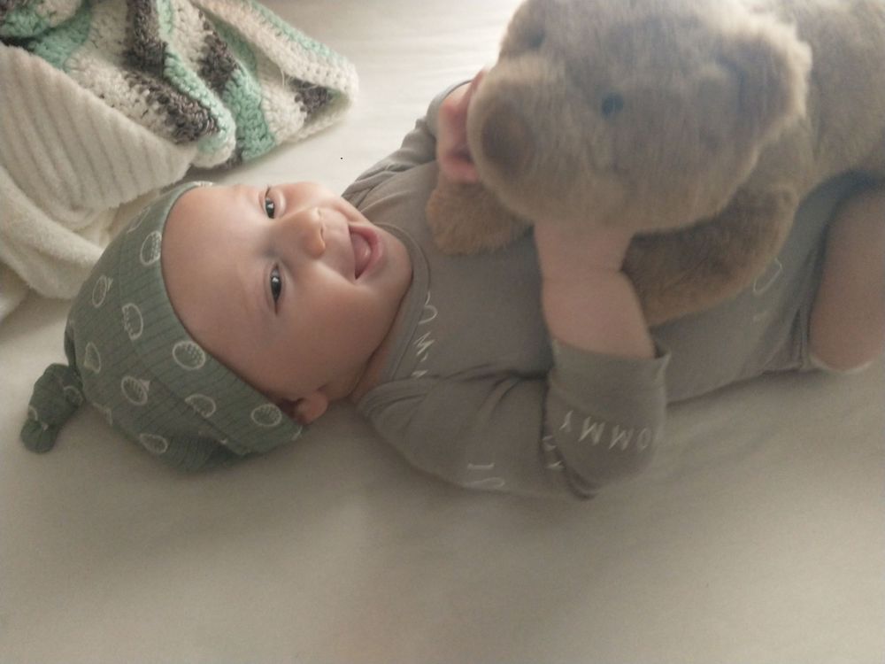 Image of a baby with a hat on laughing while holding a teddy bear.