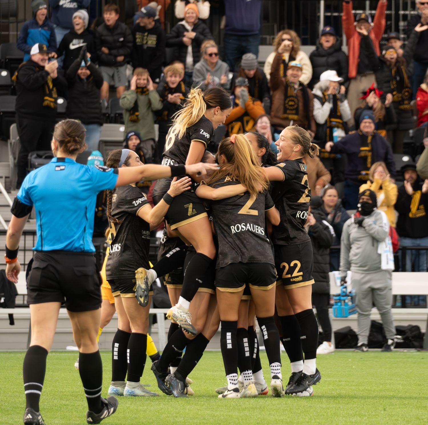 A group of athletes in black uniforms celebrate together on a sports field while an official in blue looks on.