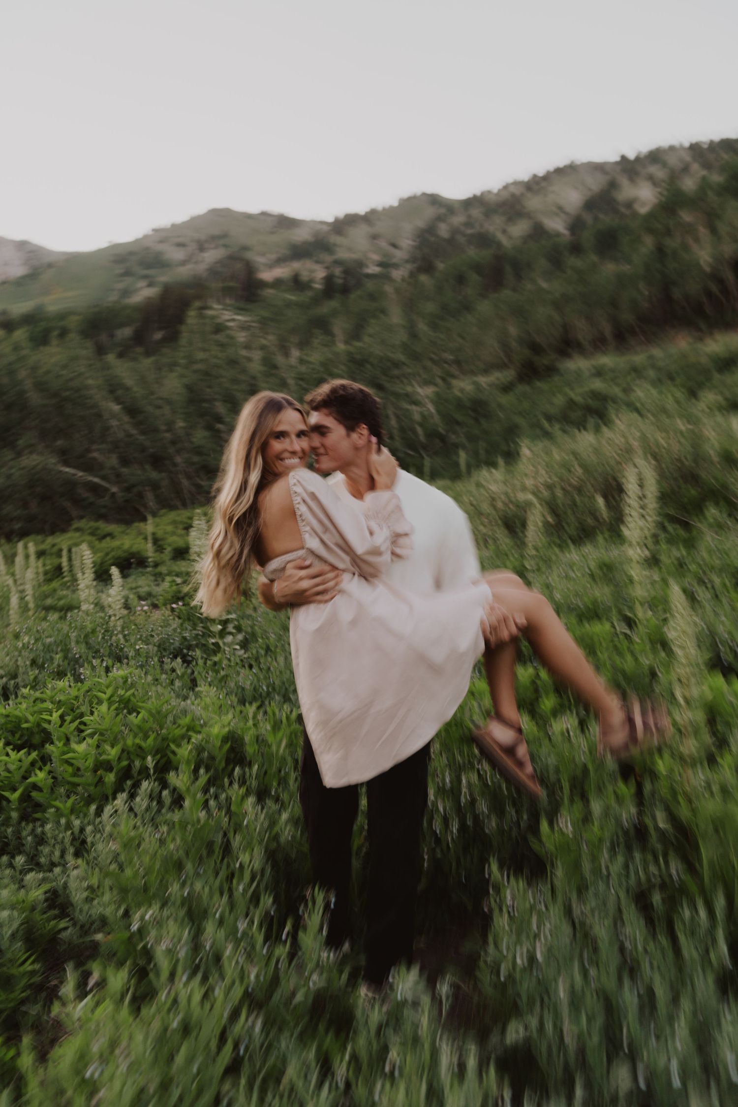 Playful couple spinning in mountain meadow with flowing white dress.