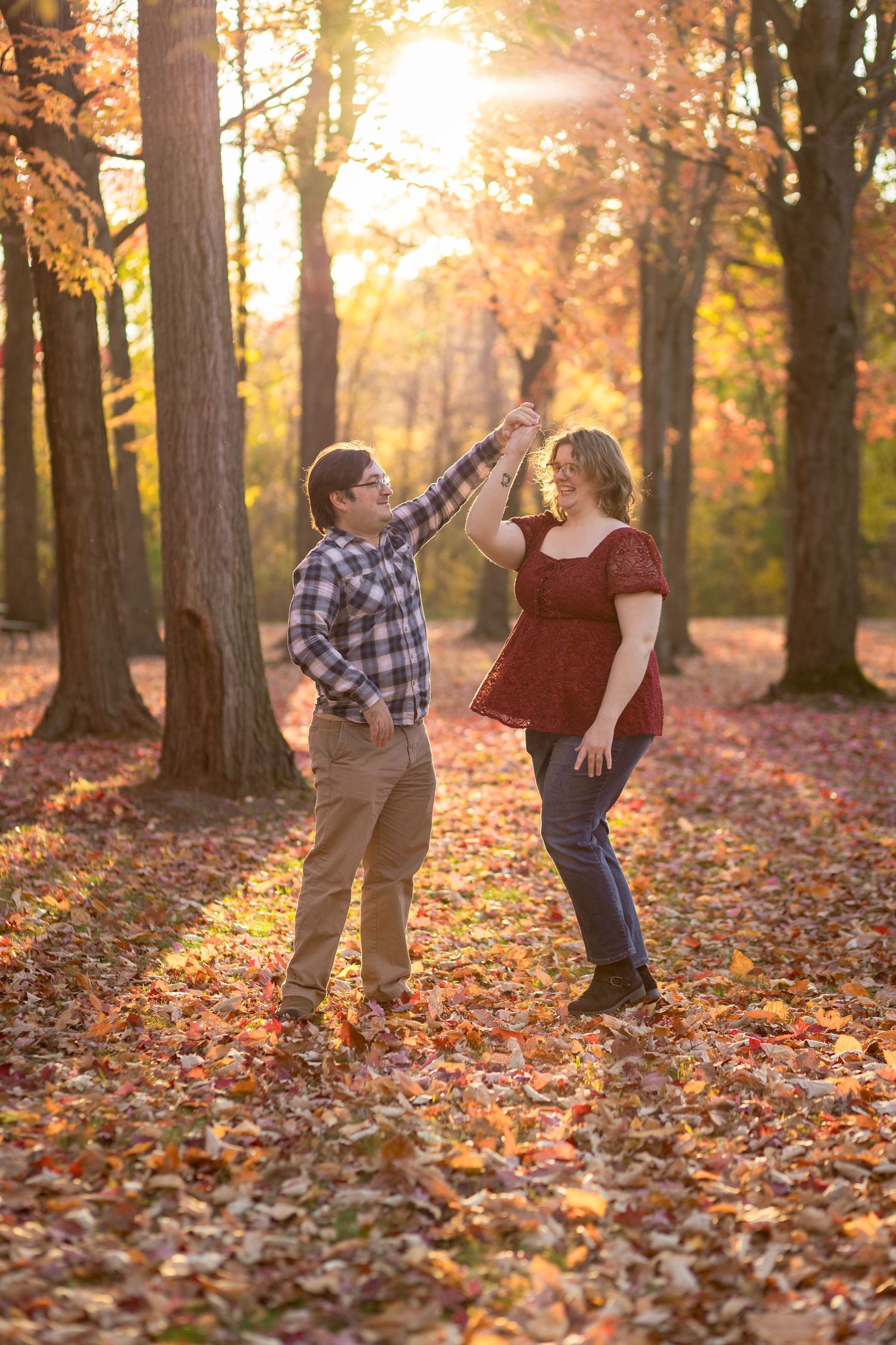 A couple playfully throws autumn leaves in a sunlit forest during a fall photoshoot.