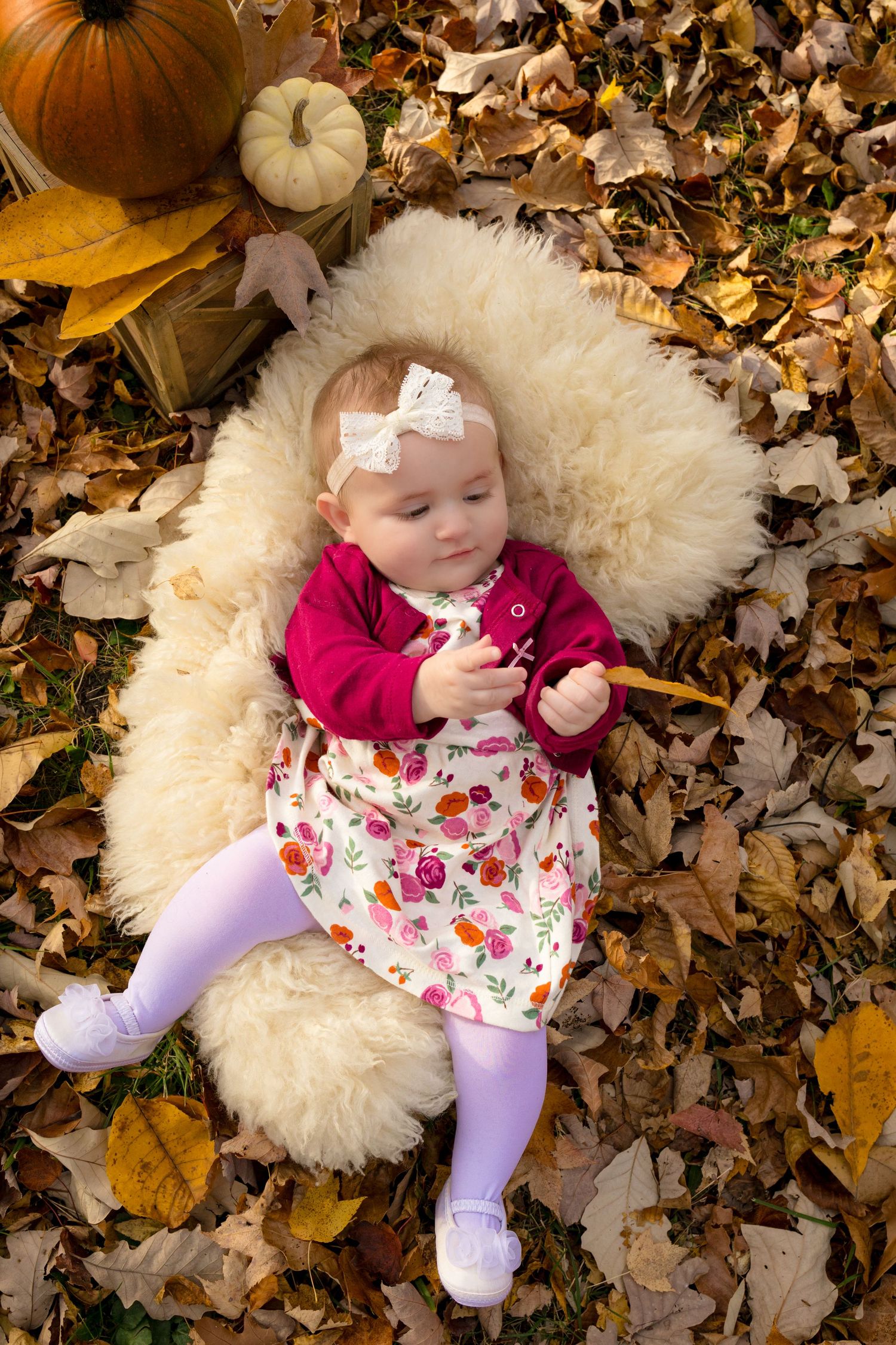 A small baby wearing a floral dress and burgundy cardigan lies on autumn leaves with a white fluffy blanket.